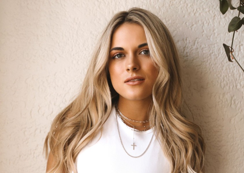A blonde woman wearing a white vest stood in front of a beige wallx